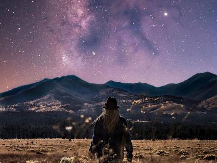 A woman stands in a field with her back to the camera looking out at mountains in Flagstaff under the Milky Way