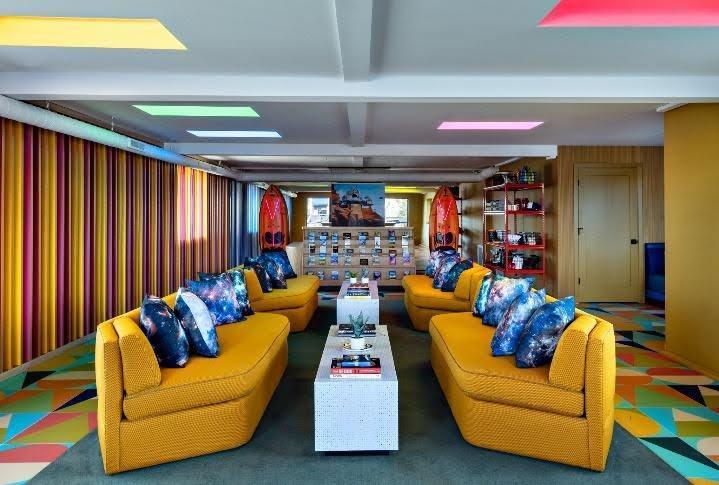 Yellow sofas are placed throughout a 70s-themed lounge, with colorful lights illuminated from the ceiling.