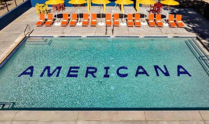 Americana Motor Hotel logo is displayed as blue tiles in a pool, beside orange lounge chairs and yellow umbrellas.