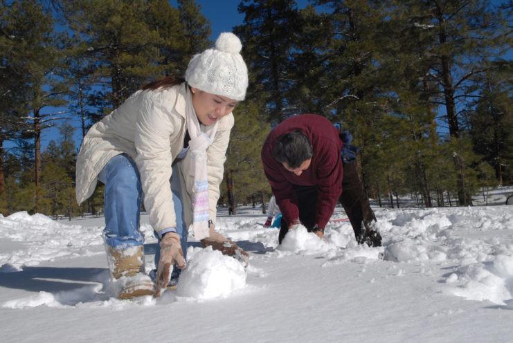 A woman in a white jacket and hat and a man in a crimson sweater in the snow rolling snowballs in Flagstaff, Arizona.