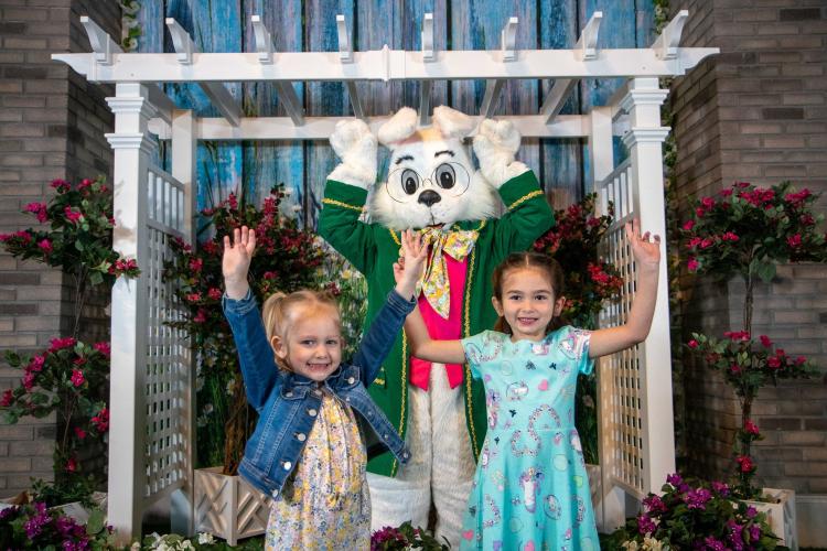Two children raise their hands in the air and pose for a photo with the Easter Bunny