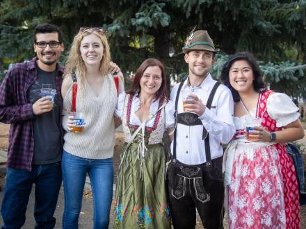 A group of people, three of which are in traditional German dress, hold cups of beer at an Oktoberfest celebration.