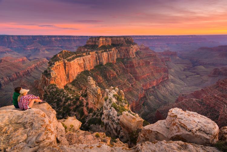 Two people sit on a boulder and gaze out at the Grand Canyon at sunset. The sky is purple and orange
