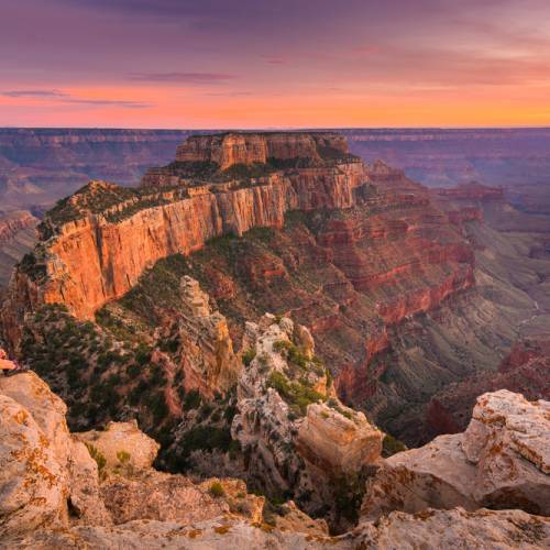Two people sit on a boulder and gaze out at the Grand Canyon at sunset. The sky is purple and orange