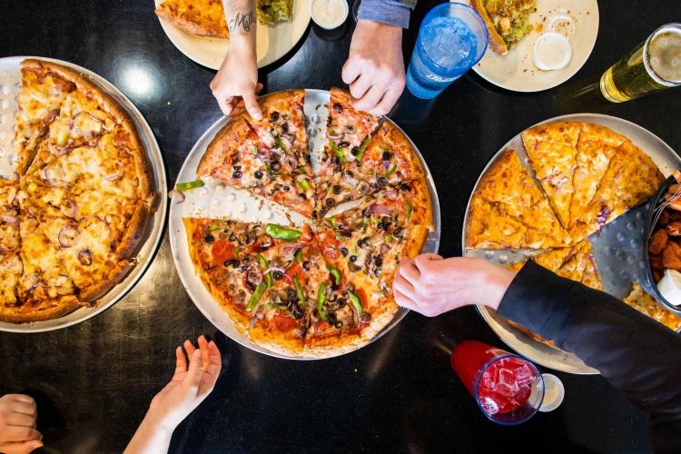 A table full of pizzas and food is surrounded by hungry patrons at NiMarco's Pizza