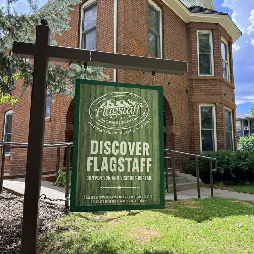 Green Discover Flagstaff sign outside on a green lawn with an old brick house in the background
