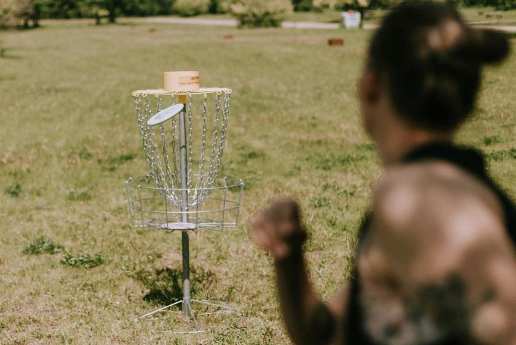 A woman, blurred, throws a frisbee into a basket in a park. 